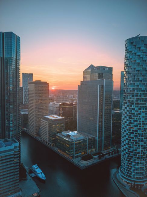 A high-angle view of a city skyline at sunset, showcasing various modern skyscrapers with reflective glass facades in shades of blue and grey. The buildings are situated along a waterway, with a white