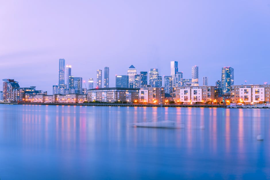 A high-angle view of a city skyline at sunset, showcasing various modern skyscrapers with reflective glass facades in shades of blue and grey. The buildings are situated along a waterway, with a white