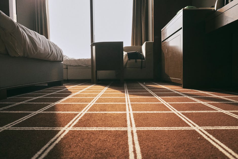 A professional cleaner dressed in white protective gear, including a hood, mask, and gloves, is performing deep cleaning and sanitisation of a light-colored carpet in a bright, minimalistic living roo