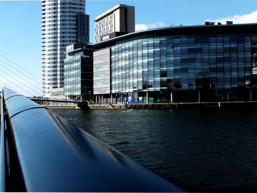 View of modern residential buildings with yellow and beige brick facades, featuring glass balconies and black window frames, lining both sides of a calm canal reflecting the blue sky and scattered clo